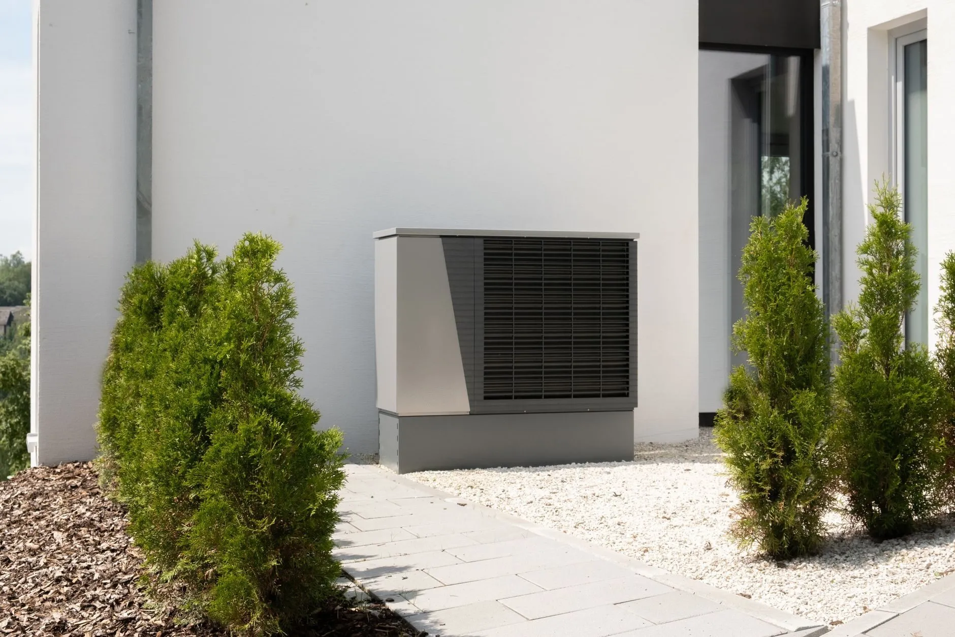 A heat pump beside a paved garden path between two conifers